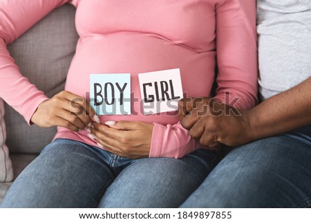 Gender reveal concept. Unrecognizable black pregnant couple holding pink and blue color card for baby boy or girl, closeup. African husband and wife guessing baby gender, sitting on couch at home