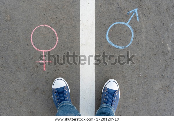 Gender symbol of a man and a woman drawn in\
chalk on the asphalt. A man\'s feet stand on the road markings\
between the gender symbols of a man and a\
woman