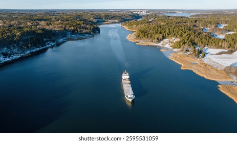 General cargo ship navigating through narrow Finnish archipelago fairway. Vessel passing near shoreline. Clear winter evening. Visible exhaust from funnel. High altitude aerial front view.