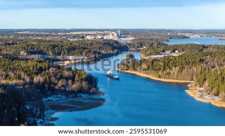 General cargo ship navigating through narrow Finnish archipelago fairway. Vessel passing near shoreline. Clear winter evening. Visible exhaust from funnel. High altitude aerial front view.