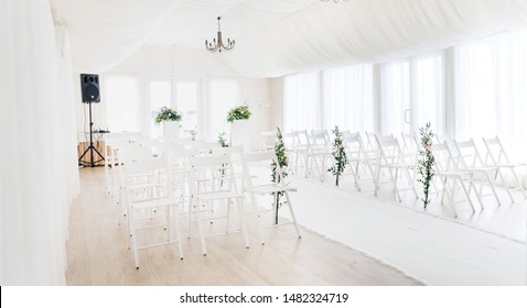 General Plan Of The Room On Wide Angle. Indoor Wedding Ceremony With White Wedding Arch Decorated With Flowers And Big White Candles.