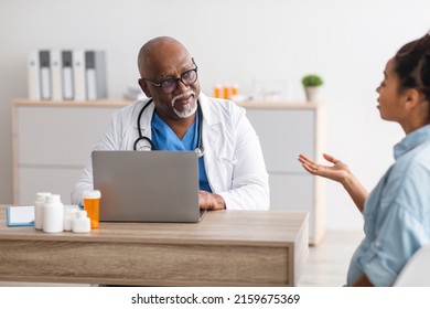 General Practitioner Appointment. Portrait of senior black male doctor in glasses and white coay listening to talking patient, sitting at table using pc. Mature specialist giving consultation to woman