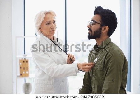 General practitioner examining heartbeat while providing regular checkup in clinic. Side view of focused elderly lady in white coat using stethoscope while young man sitting on exam couch.