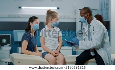 General practitioner with face mask taking notes on clipboard files and consulting small girl in cabinet. Medic using checkup documents to give prescription treatment and medicine.