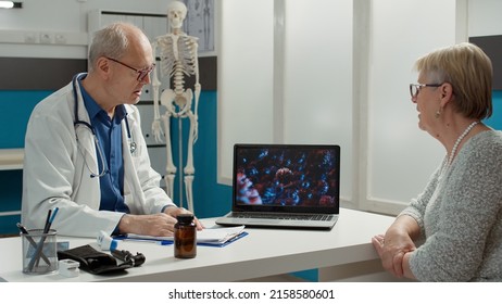 General Practitioner And Patient Analyzing Covid 19 Virus Animation On Laptop, Talking About Prevention And Recovery In Medical Office. Doctor Using Coronavirus Illustration On Screen.