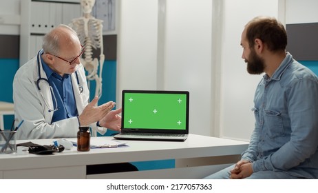 General Practitioner Pointing At Laptop Display With Greenscreen, Meeting At Checkup Visit. Looking At Blank Copyspace Background With Isolated Mockup And Chroma Key Template. Tripod Shot.