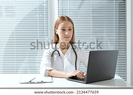 General practitioner working on laptop while she sitting behind table in private clinic or hospital. Doctor fills update info on patient record, make treatment plan, reviews schedule of appointments