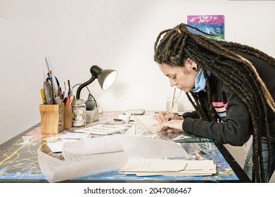 General Shot Of Beautiful Young White Latin Hispanic Art Director Girl, With Dreadlocks And Good Style, In Her Production Studio, Tracing A Shot On Onion Paper, Working On Her Well Lighted Work Table.