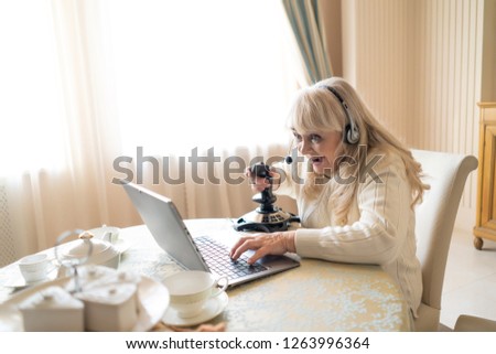 General Shot Of Excited Senior Woman Winning In Video Game Holding A Joystick And Wearing Headphone. Sitting At The Table Playing Games