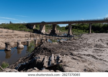 General view of bridge construction site showing pile foundations, stream channel, and existing bridge background