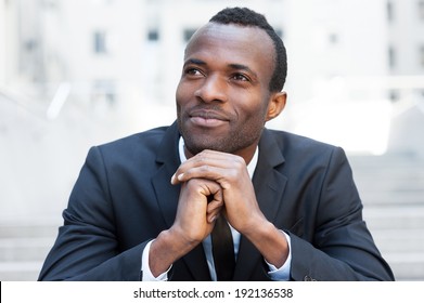 Generating new great ideas. Cheerful young African man in formal wear keeping hands clasped and looking away while sitting at the outdoors staircase