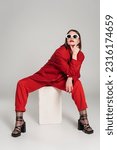 generation z, fashion model with brunette short hair and nose piercing posing in sunglasses and red suit while sitting on concrete cube on grey background, lady in red, young woman