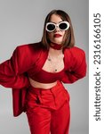 generation z, tattooed young woman with brunette short hair and nose piercing posing in sunglasses and red suit on grey background, modern fashion, trendy outfit, chic style