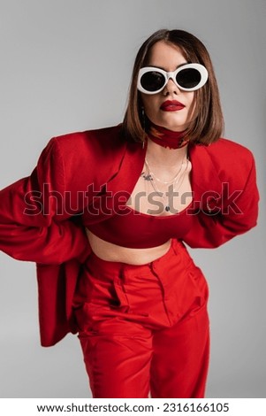 generation z, tattooed young woman with brunette short hair and nose piercing posing in sunglasses and red suit on grey background, modern fashion, trendy outfit, chic style