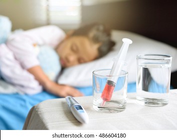 Generic Liquid Medicine Syringe In Glass On Table In Front Of Out Of Focus Sleeping Child For Health And Illness Concept, Young Asian Girl Hugs The Blue Teddy Bear.