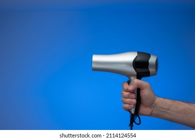 Generic Silver Hair Dryer Held In Hand By Caucasian Male Arm. Close Up Studio Shot, Isolated On Blue Background, No People.