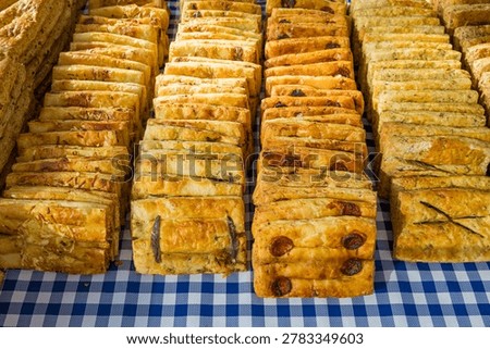 A generous and appetizing photograph of multiple rows of golden savory pastry slices displayed for sale on a blue and white checkered tablecloth at an outdoor French market. The baked goods are arranged in neat overlapping rows, each variety distinguished by its visible filling and topping. From left to right the varieties include a cheese and herb loaf cake, an olive and herb fougasse-style slice, a chorizo and tomato pastry, and a rosemary and herb-seasoned variety. All are baked to a rich golden-brown and display a moist, dense crumb with visible ingredient inclusions. The abundant display and rustic presentation evoke the atmosphere of a traditional French farmers market or village fair. The warm, inviting tones of the baked goods against the classic blue and white gingham cloth reinforce the authentic, artisan character of the scene. This image suits French food culture, artisan bakery and delicatessen marketing, farmers market and local produce content, food tourism, gastronomy, recipe and culinary editorial, hospitality, and southern France regional lifestyle campaigns.
