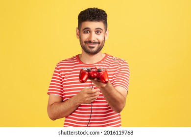 Generous bearded man in striped t-shirt holding in hand gaming joystick, suggesting you to play video game with him, virtual reality simulator. Indoor studio shot isolated on yellow background