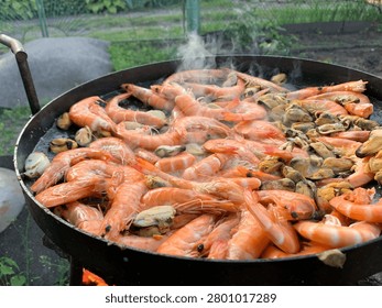 A generous portion of succulent shrimp and briny mussels are sizzling in a large black pan, releasing aromatic steam as they cook over a vibrant flame
