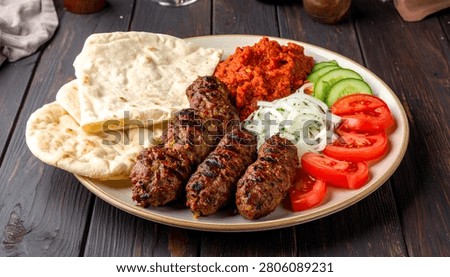 A generous serving of grilled cevapi, served with warm flatbread, a vibrant red pepper spread, creamy yogurt dip, and sliced tomatoes and cucumbers.