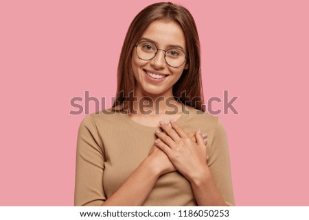 Generous thankful friendly looking girl keeps hands on heart, has good attitude, dressed in casual clothes, isolated over pink background. Joyful king lady feels thankfulness. Horizontal view