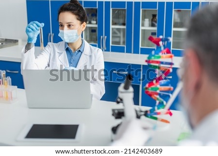 geneticist in medical mask working with test tube and laptop near blurred colleague