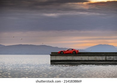 Geneva, Switzerland - March 2020: Red Ferrari F12 TdF Italian rosso supercar parked on pier by Lake Geneva (Lac Léman) during sunset  with Swiss alps in the background