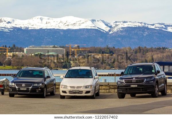 Geneva, Switzerland - March 9, 2018: Three black and\
white empty cars parked on parking lot on background of lake water,\
woody urban landscape and mountain range with snowy peaks under\
sunny blue sky.