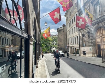 Geneva, Switzerland - September 15 2021: A Cyclist Is Riding On A Small Old Town Street With National And City Flags Around And Reflection In A Store Window