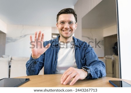 A genial man waves at the camera, with a tablet and laptop on the table, in a modern home environment.