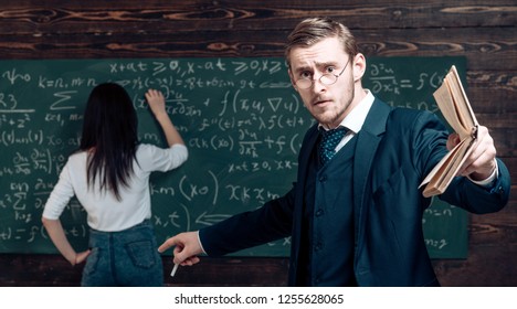 Genius Man In Glasses With Book Point At Woman Solving Equation On Chalkboard. Genius Teacher With Student In Classroom.