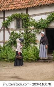 Genk, Belgium - August 11, 2021: Domein Bokrijk. Both In Traditional 19th Century Clothing, Female Doctor Visits Lady Of Farm Meeting Her Outside White Painted House With Red Roof. Green Foliage.