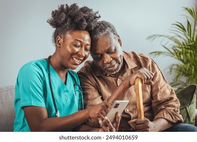 A gentle Black mid-adult nurse aids a senior male with a smartphone, both engaged in the activity. They are comfortably seated in a home setting, reflecting professional in-home healthcare. - Powered by Shutterstock