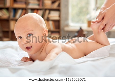 Gentle and careful young adult mom making feet massage to her little new born baby while child lying in bedroom on cozy bed