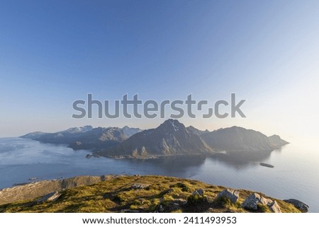 Gentle hues of the midnight sun illuminate the Lofoten Islands, as seen from Offersoykammen, highlighting the dramatic interplay of mountain and ocean