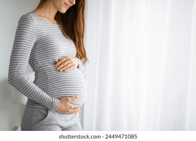 A gentle image capturing a pregnant woman in casual clothes, tenderly holding her belly, standing by a window with white curtains - Powered by Shutterstock