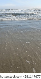 Gentle Layers of Ocean Waves Approaching Shore. A wide vertical landscape displays soft ripples of water on a wet sandy beach leading toward multiple layers of white cresting waves under a pale blue sky.
