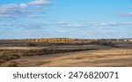 Gentle rolling hills of harvested fields extend towards a vibrant treeline alight with the fiery colors of fall, under a vast sky with wispy clouds.