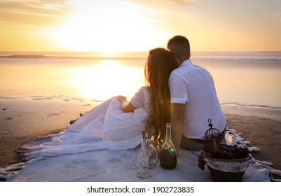 A gentle romantic date on the ocean during the magical sunset. A girl and a guy in white clothes sit on a white bedspread on the ocean during a magical sunset.