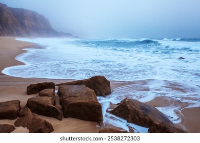 Gentle waves break over large rocks along a misty shoreline, creating frothy patterns in the sand under a calm, overcast sky - Powered by Shutterstock