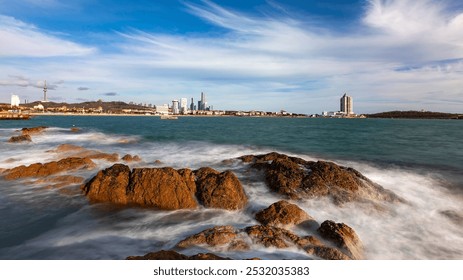 Gentle waves crash over rocky shoreline; distant cityscape silhouettes under a sky with wispy clouds, creating a serene coastal scene. - Powered by Shutterstock