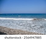 Gentle waves roll onto a pebbly beach under a clear blue sky during daytime. The calm sea stretches into the horizon, inviting relaxation and enjoyment of natures beauty.