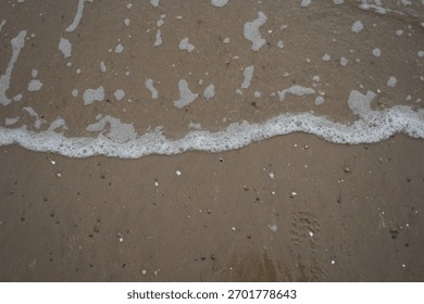 Gentle white foam from the ocean waves washes across the wet, textured brown sand of Llanddwyn Beach, Anglesey, North Wales, revealing small scattered pebbles and shells on the shore.