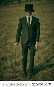 A gentleman in a black suit is standing in a field with a top hat pulled over his eyes to hide his face. Hiding from reality. Surrealism.