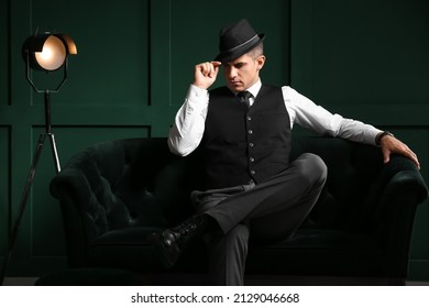 Gentleman in formal clothes and stylish hat sitting on sofa against color wall