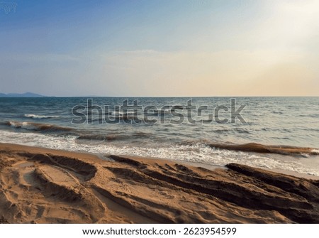 A gently sloping beach with small waves rolling onto the shore under a soft, glowing sky. The sunlight casts a warm tone across the water and sky, creating a tranquil, late-afternoon ambiance. 