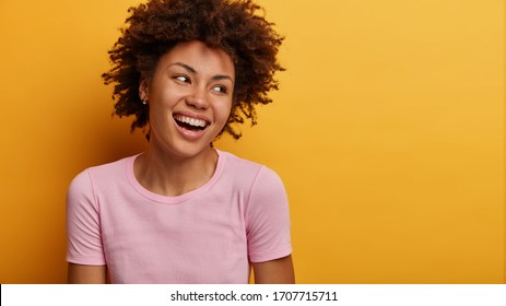 Genuine Emotions, Happiness Concept. Positive Dark Skinned Woman Laughs Out From Hilarious Joke, Concentrated Aside, Cannot Hold Laughter As Talks With Funny Friend, Isolated On Yellow Background