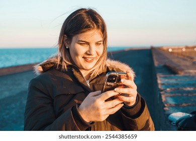 Genuine happy young woman using smartphone at seaside pier during sunset wearing winter jacket enjoying social media connection - Powered by Shutterstock