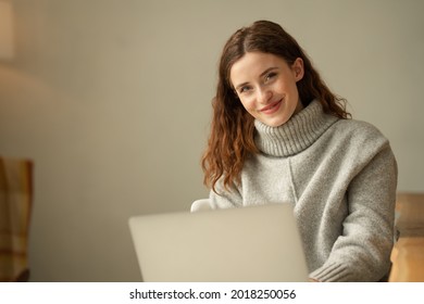 Genuine Young Woman With A Sweet Friendly Smile And Attentive Expression Working On A Laptop At Home During The Covid-19 Pandemic Looking At The Camera