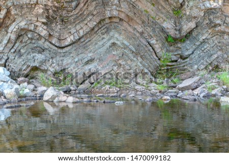 A geological fold in sedimentary rock. The fold is in a cliff above a river. Many layers of sedimentary rock visible. Plants grow from the rock.